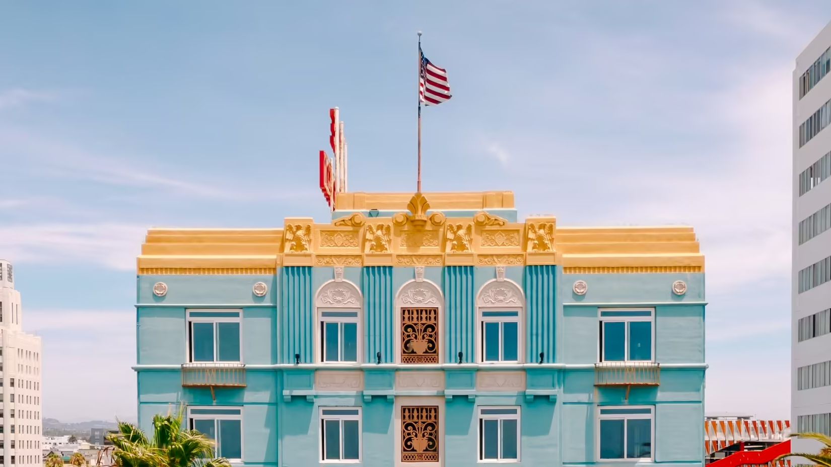 A light blue ornate building with gold trim, flags on top, and a clear sky behind it, framed by surrounding taller buildings.