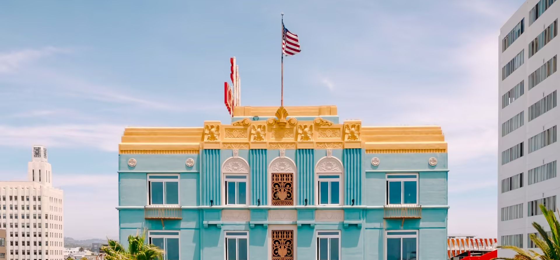 A light blue ornate building with gold trim, flags on top, and a clear sky behind it, framed by surrounding taller buildings.