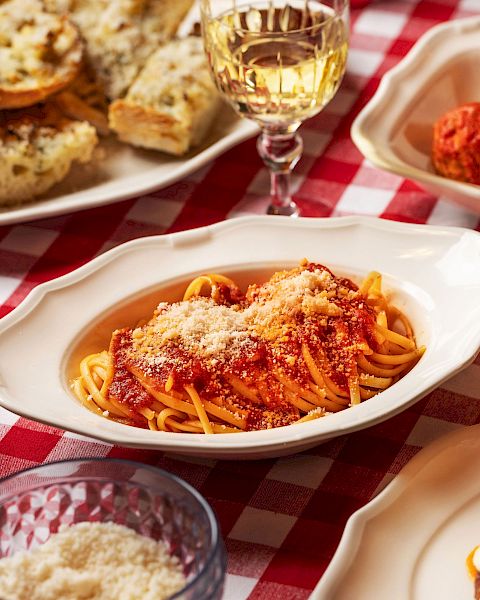 A table with Italian dishes: pasta in tomato sauce, garlic bread, meatballs, salad, and a glass of white wine on a red-checkered tablecloth.