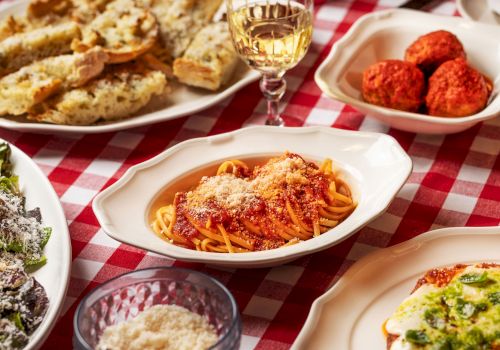 A table with Italian dishes: pasta in tomato sauce, garlic bread, meatballs, salad, and a glass of white wine on a red-checkered tablecloth.