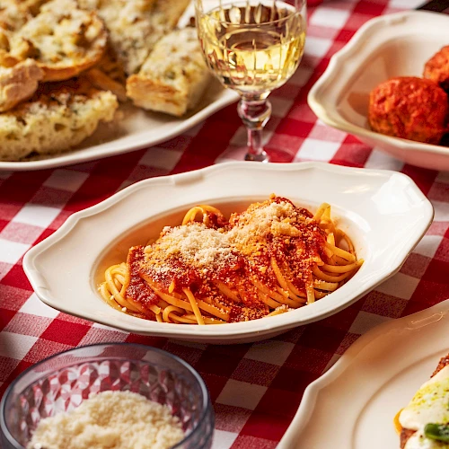 A table with Italian dishes: pasta in tomato sauce, garlic bread, meatballs, salad, and a glass of white wine on a red-checkered tablecloth.