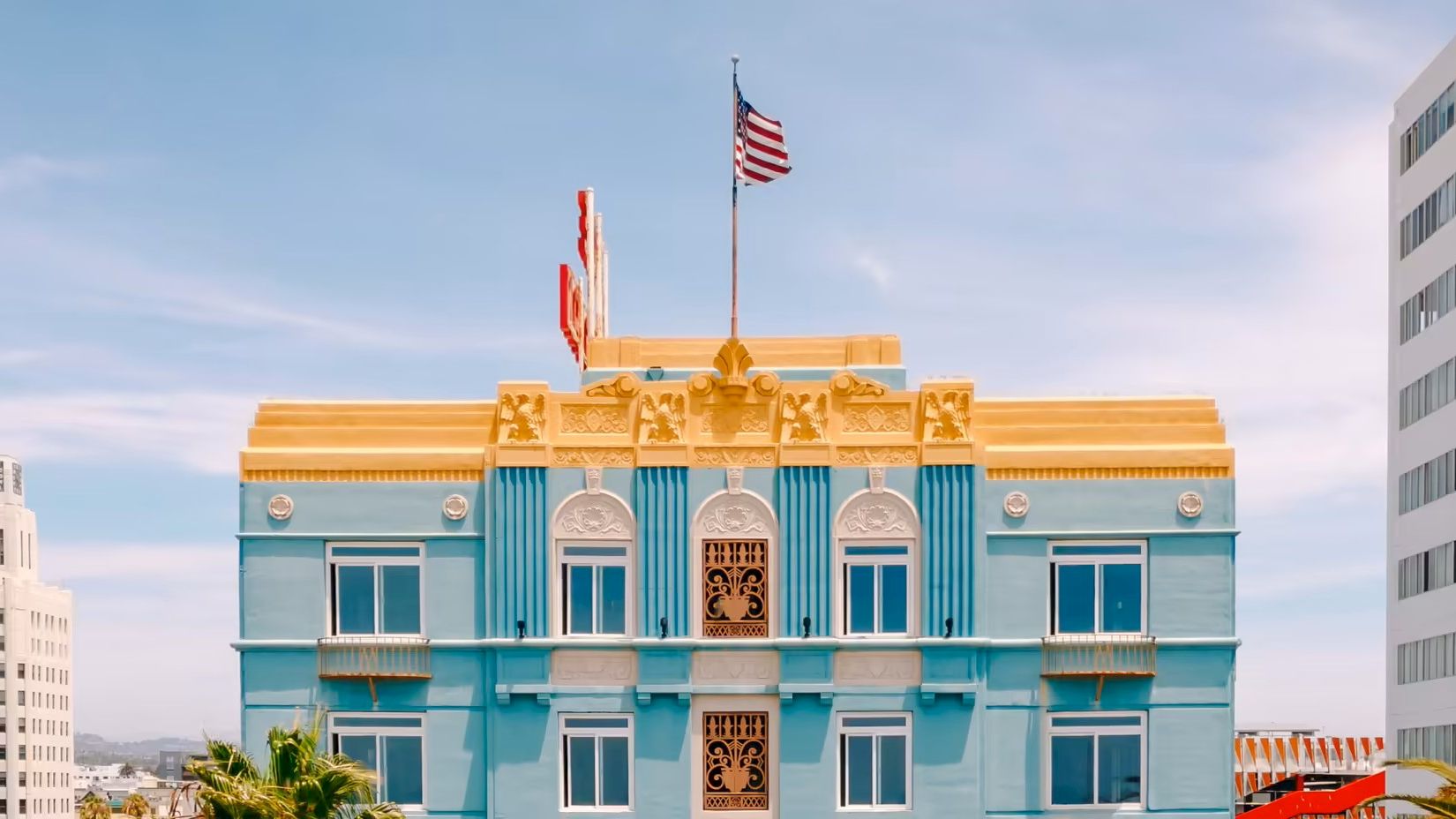 A pastel blue, ornate building with gold trim and arched windows, flagpole, and an American flag atop; surrounding cityscape.