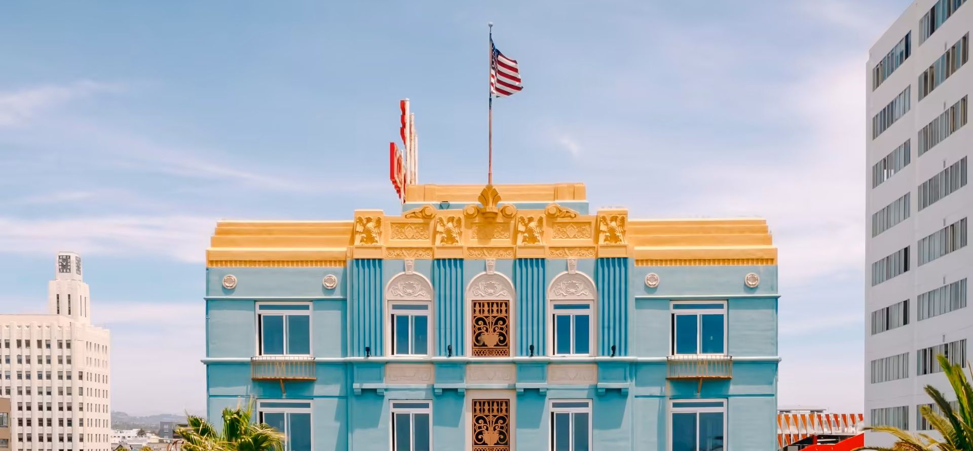A pastel blue, ornate building with gold trim and arched windows, flagpole, and an American flag atop; surrounding cityscape.