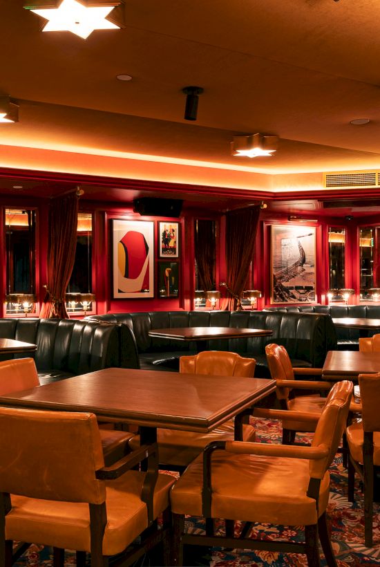 A cozy, retro-style dining lounge with tan chairs, wooden tables, red curtains, framed art, and warm ambient lighting.