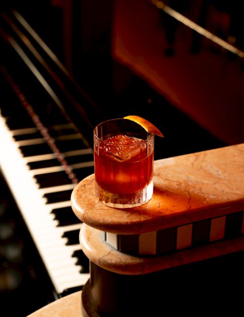 A cocktail with a citrus garnish sits on a carved wooden pedestal beside a piano, warmly lit and inviting.