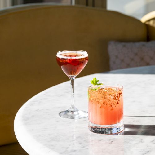 Two cocktails on a marble table by a window, one red in a coupe and one pinkish with mint garnish.