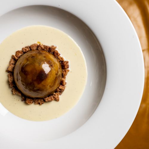 A fancy plated dessert: a glossy domed pastry on a pale sauce with a ring of crunchy crumble around it, served on a white plate.