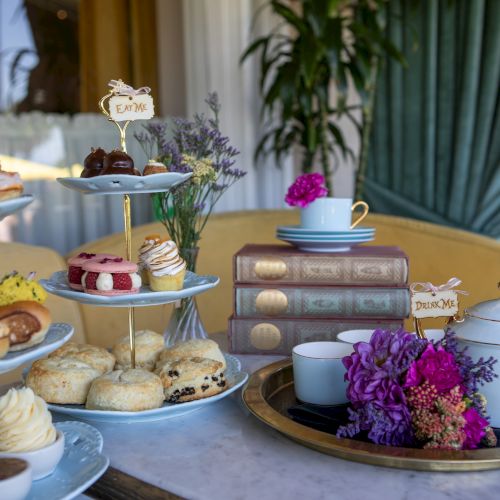 A three-tier stand with assorted pastries, scones, and cakes beside a teacup set, bowls, flowers, and a teapot on a cozy, elegant table.