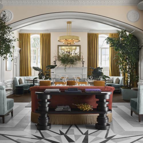A grand, elegant hotel lobby with tall gold curtains, oversized plants, plush seating, a central table, and a starburst floor design.