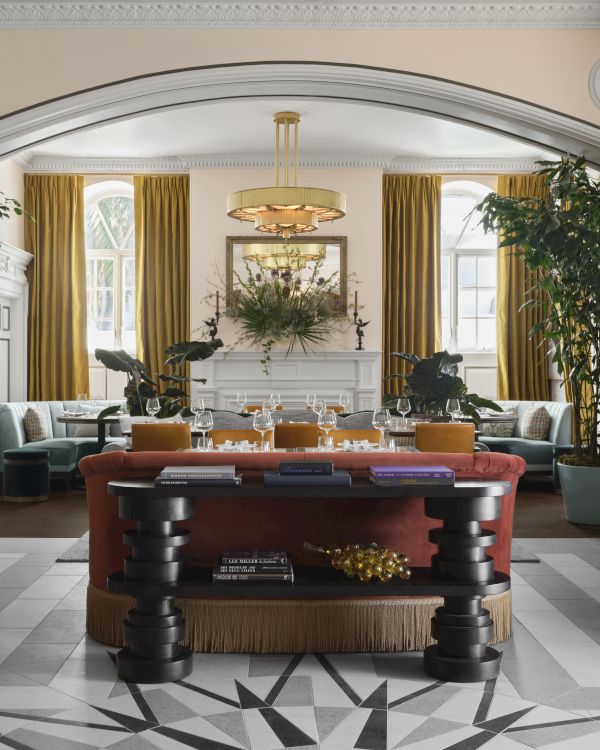 A grand, elegant hotel lobby with tall gold curtains, oversized plants, plush seating, a central table, and a starburst floor design.