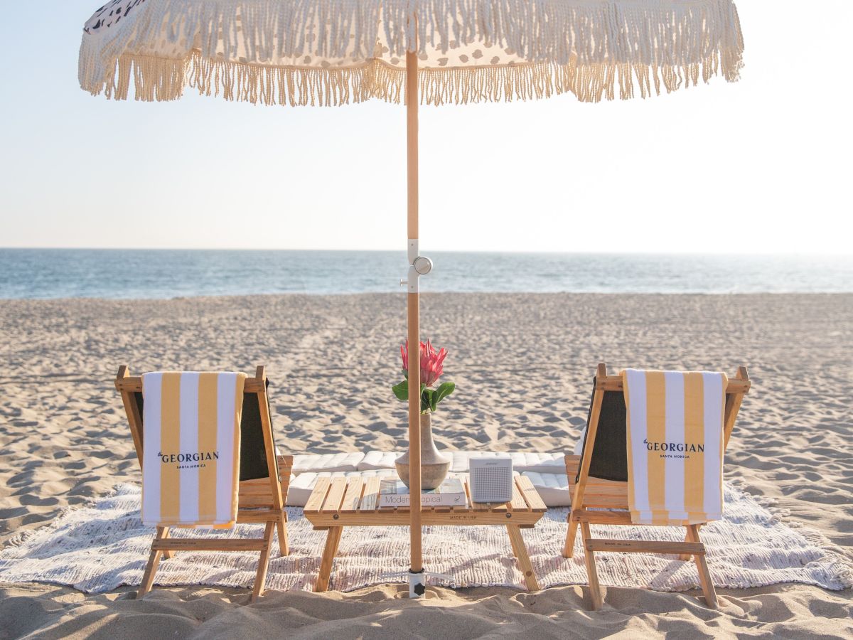 Two striped beach chairs face the ocean under a thatched umbrella, with a small wooden table between them holding a vase of flowers on a sunny, sandy shore.