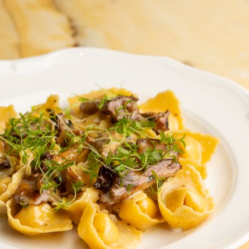 A plate of pasta topped with sliced meat, greens, and a light sauce on a white dish, set on a marble table.