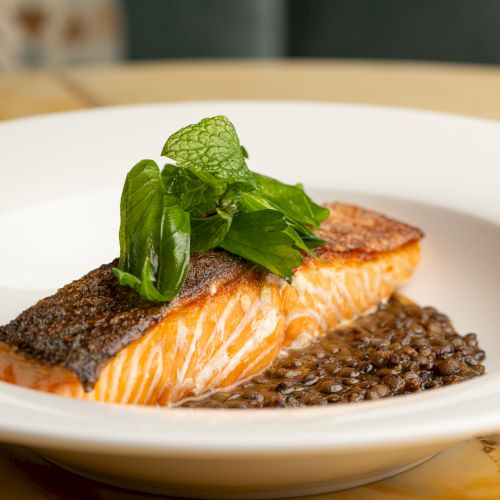 A plated salmon fillet with crispy skin, garnished with fresh mint and basil, resting on a bed of lentil or grain puree on a white plate. Ending sentence.