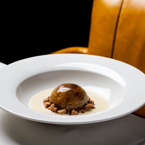A small, polished dish with a tiny dark garnish or sauce center on a white plate, set on a light table with a wooden background.
