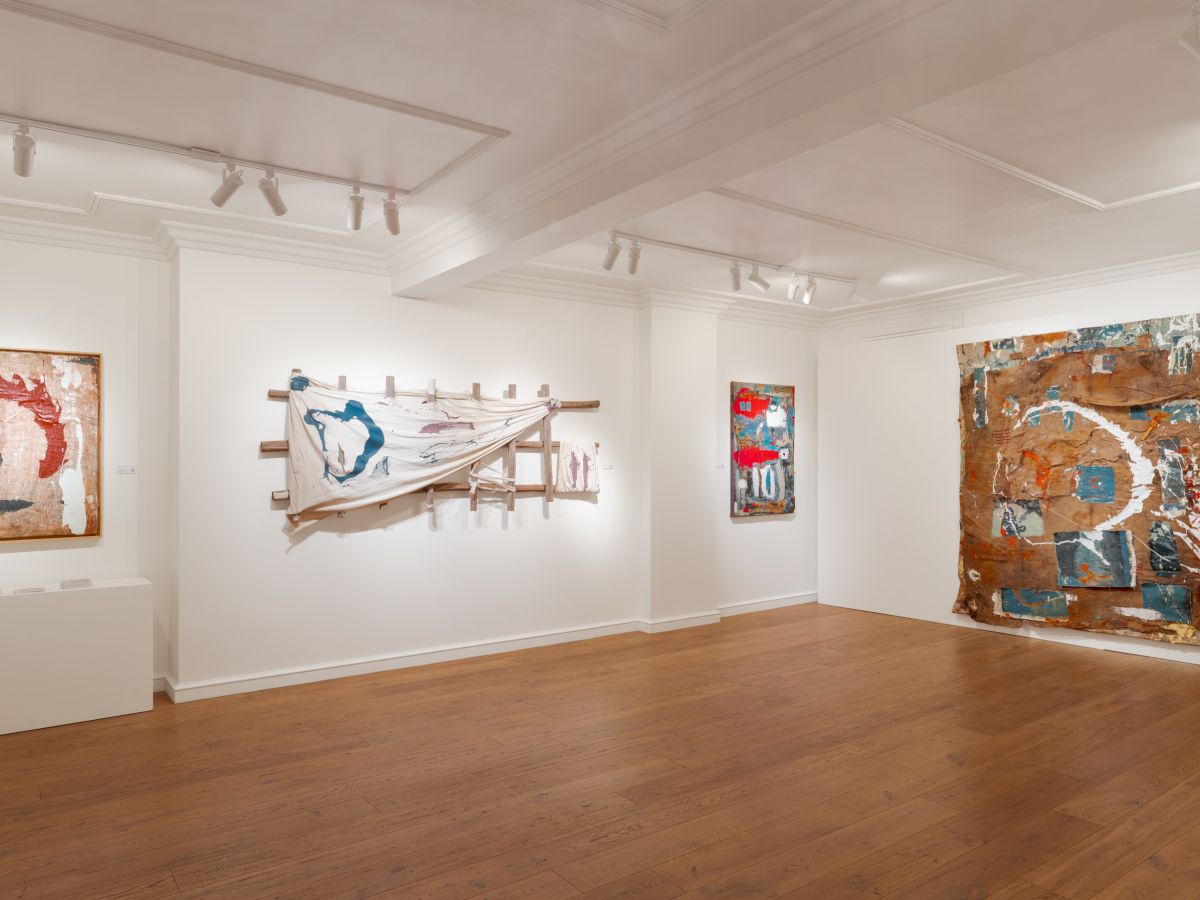 An airy gallery with white walls and wooden floors, displaying abstract, mixed-media artworks on the walls and a raised sculpture in the center, all under soft track lighting.