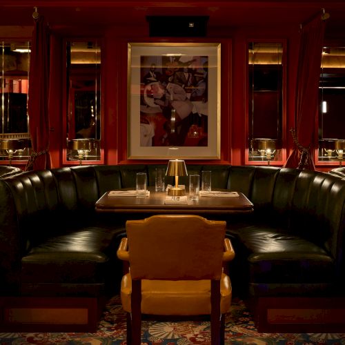 A cozy dining booth with a black leather U-shaped sofa, a wooden chair, a framed poster, and a candle-lit table in an elegant, dimly lit restaurant.