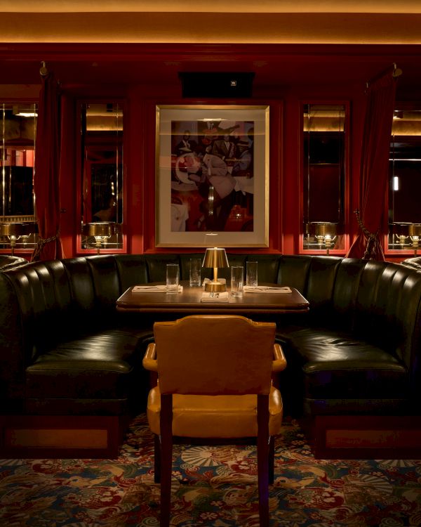 A cozy dining booth with a black leather U-shaped sofa, a wooden chair, a framed poster, and a candle-lit table in an elegant, dimly lit restaurant.