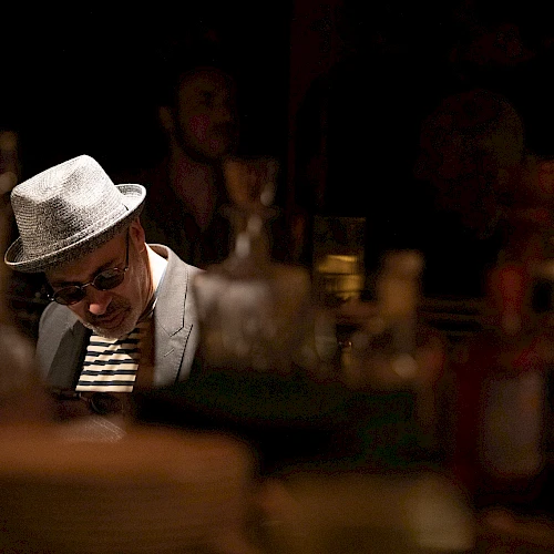 A man in a light fedora and sunglasses sits at a dim bar, looking down, with bottles and warm lights blurred in the foreground.