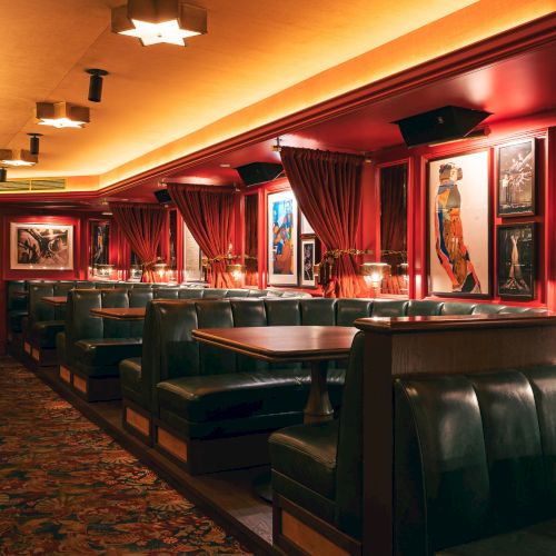 A dimly lit upscale dining area with red walls, booths, framed art, and warm yellow lighting along a carpeted floor, cozy and intimate.