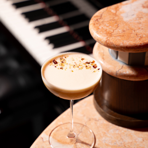 A creamy cocktail in a coupe sits beside a marble table and piano keys, with a round dessert perched nearby.