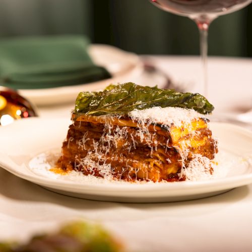 A plated lasagna-like dish topped with greens and grated cheese sits on a white plate at a fine-dining table.