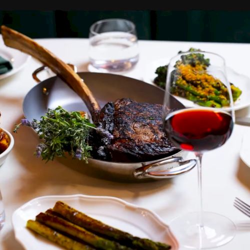 A fancy steak dinner with a large ribeye, a bread basket, roasted asparagus, a herb garnish, and a glass of red wine on a white tablecloth.