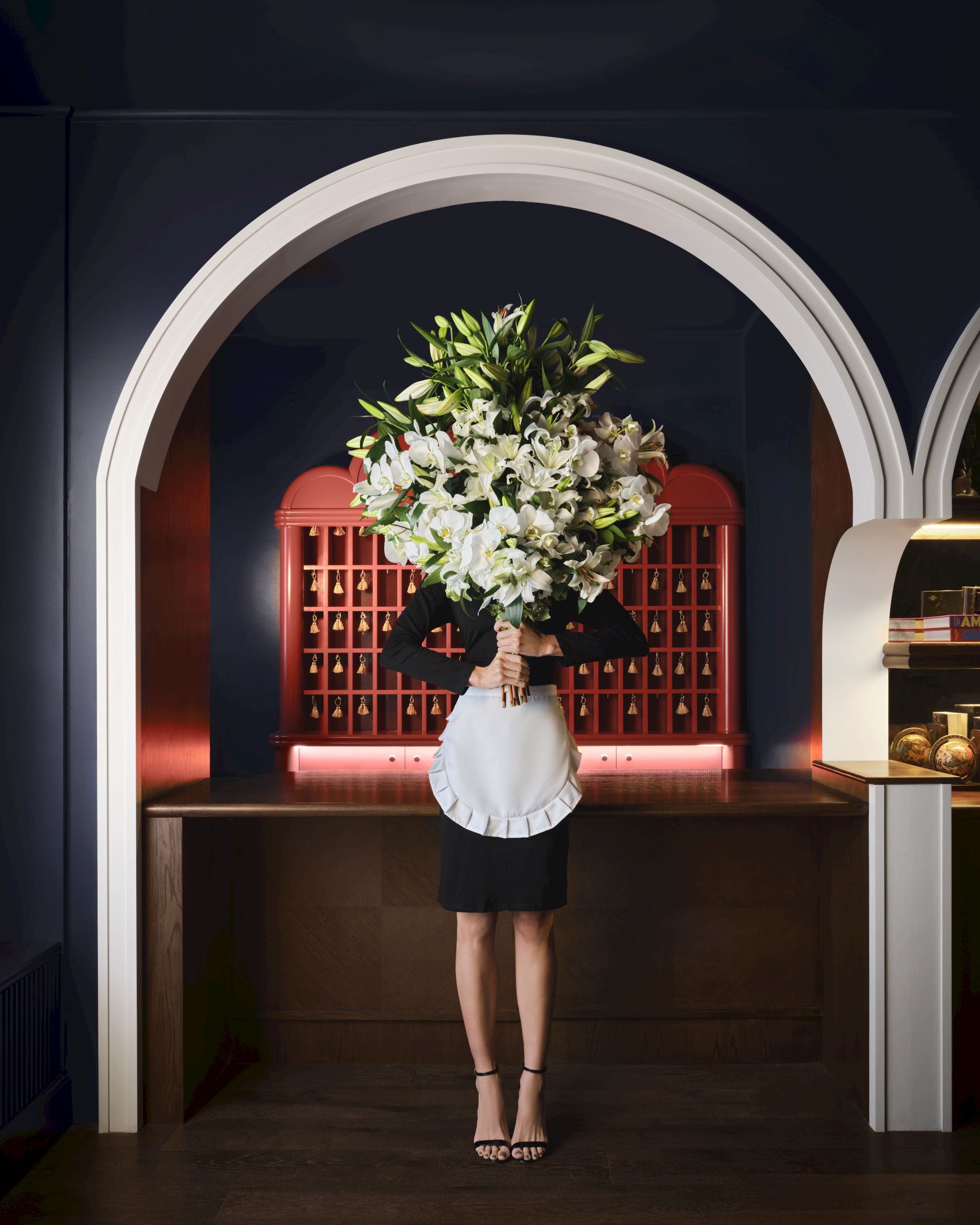A person with a large bouquet hides their face behind the flowers at a stylish, arched bar counter, wearing a white blouse and black skirt.