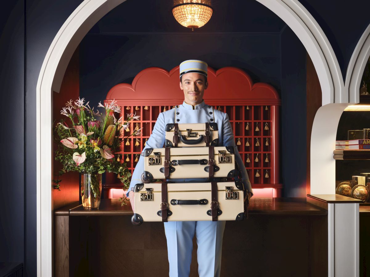 A chef dressed as a knight stands in a stylish kitchen, holding stacked suitcases like a tray, with flowers and warm lighting behind.