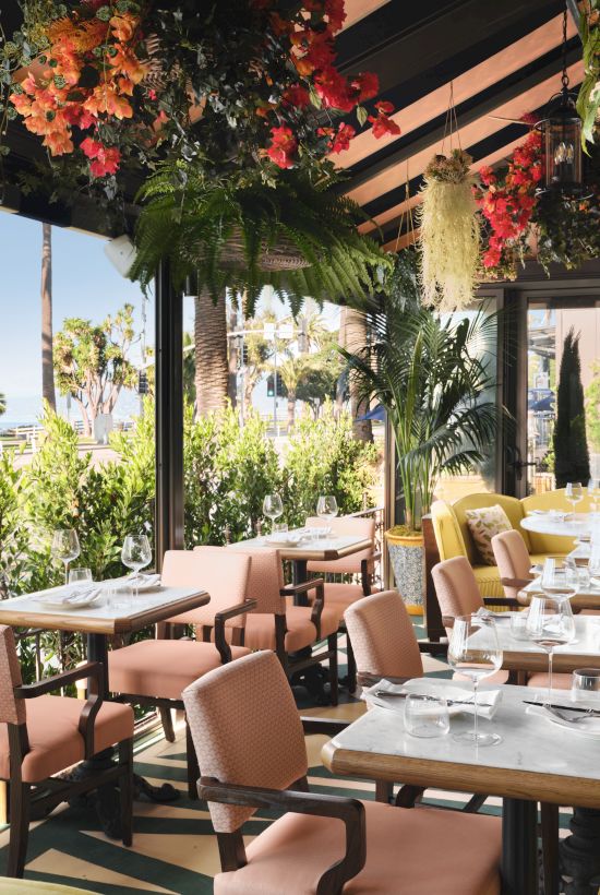 A stylish outdoor dining area with pastel pink chairs, yellow seating, and hanging greenery under a shaded terrace, set for brunch.