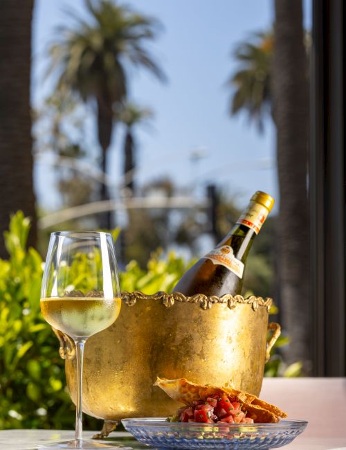 A bottle of wine in a brass bucket with a glass, a plate of food, and a sunny outdoor scene with palm trees.