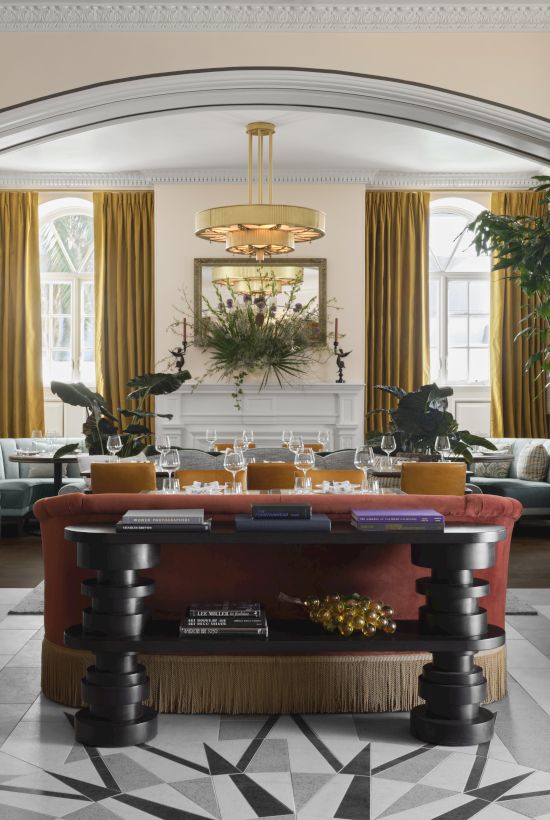 A luxurious hotel lobby with high ceilings, gold curtains, a grand centerpiece table, green plants, and stylish seating around a striking geometric floor pattern.
