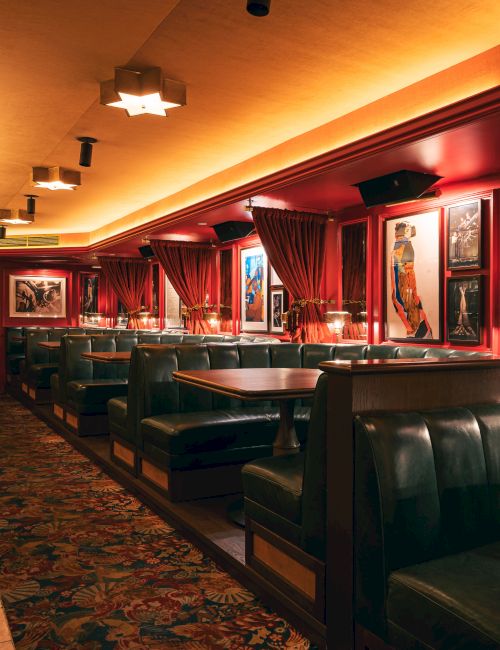 A vintage-style lounge or club with red velvet booths, framed artwork, warm ambient lighting, and a patterned carpet. 140 characters: A vintage-style lounge with red velvet booths, framed art, warm lighting, and a patterned carpet, evoking classic club ambiance.