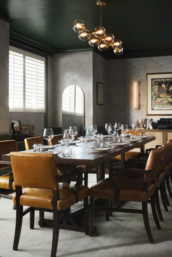 A stylish dining room with a long wooden table, tan chairs, modern chandelier, gray walls, large windows, and a sideboard with artwork.