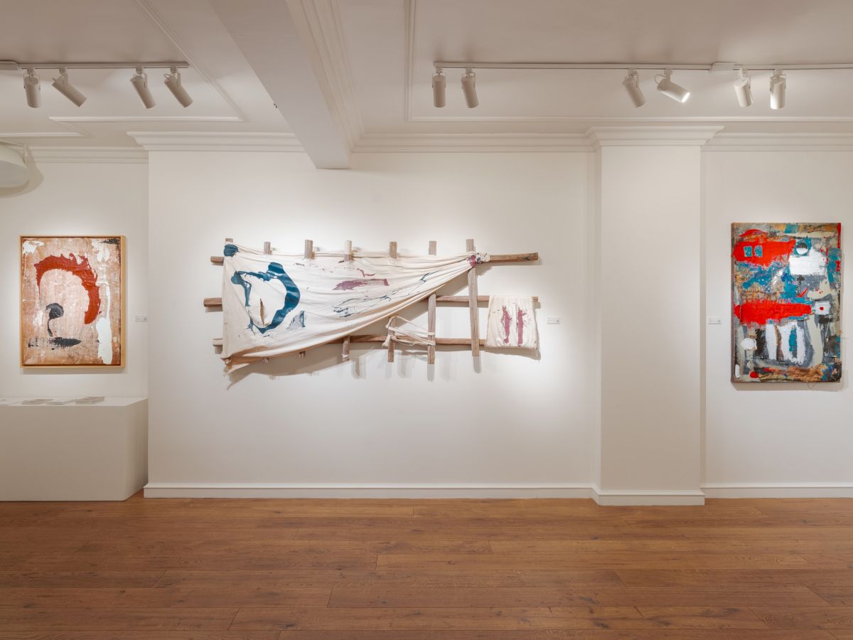 A modern art gallery with white walls, wooden floor, three abstract paintings, and a sculptural wooden installation centered between them, lit by track lights.