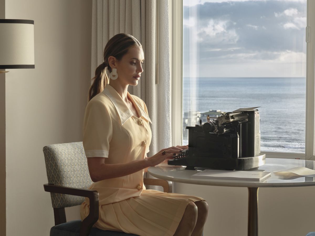 A woman sits at a small round table by a large window overlooking the ocean, typing on a vintage sewing machine in a pale-yellow dress.