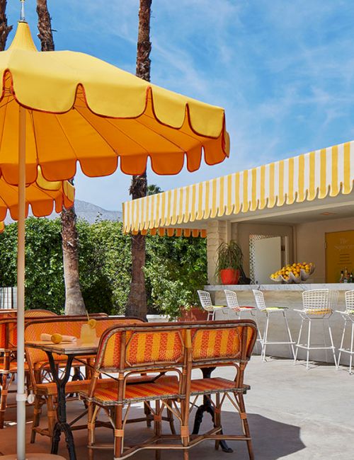Outdoor cafe scene with bright yellow umbrellas, wooden tables and chairs, a striped awning, and a bar area under sunny skies.