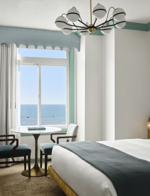 A bright hotel room with a view of the sea, featuring a yellow-accent bed, white linens, a round table by the window, and a stylish chandelier.