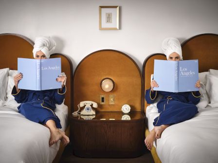 Two people in robes with towels on their heads sit in bed, reading blue books, on either side of a shared nightstand between them.