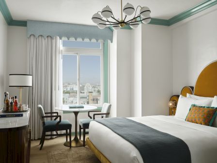 A bright hotel room with a large window, round table and chairs, a bed with a yellow headboard, and a modern chandelier.