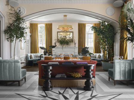A luxurious hotel lobby with a central wooden console, gold-framed chairs, tall yellow curtains, lush green plants, and a sunburst marble floor.