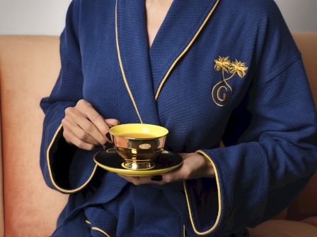 A person wearing a blue bathrobe sits on a couch, holding a small decorative teacup and saucer with gold trim.
