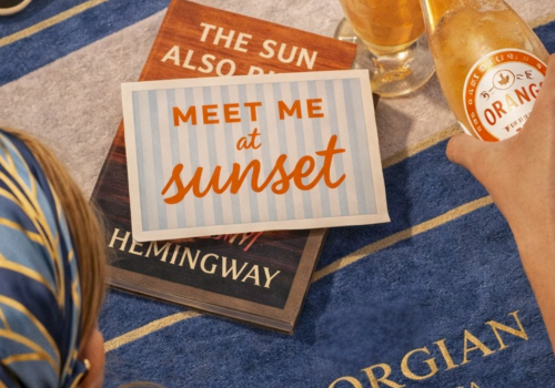 A beach scene with a blue towel, sunglasses, a bottle and glass of sparkling drink, and books labeled &ldquo;Meet me at sunset&rdquo; at sunset.
