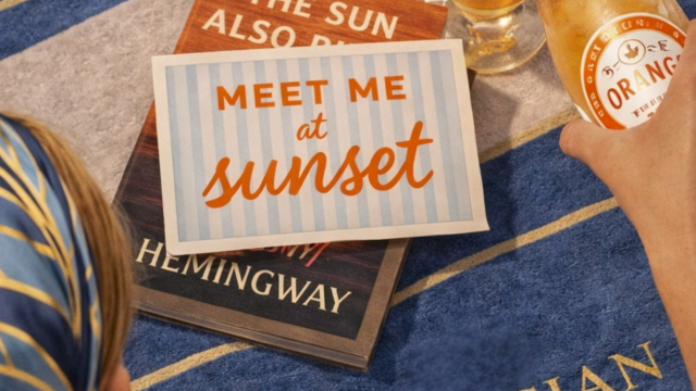A beach scene with a blue towel, sunglasses, a bottle and glass of sparkling drink, and books labeled &ldquo;Meet me at sunset&rdquo; at sunset.