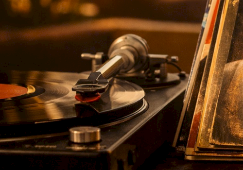 A vintage turntable records a vinyl with a tonearm playing; a spinning record, with a dusty, warm-toned close-up, on a wooden shelf.