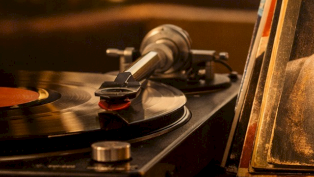 A vintage turntable records a vinyl with a tonearm playing; a spinning record, with a dusty, warm-toned close-up, on a wooden shelf.