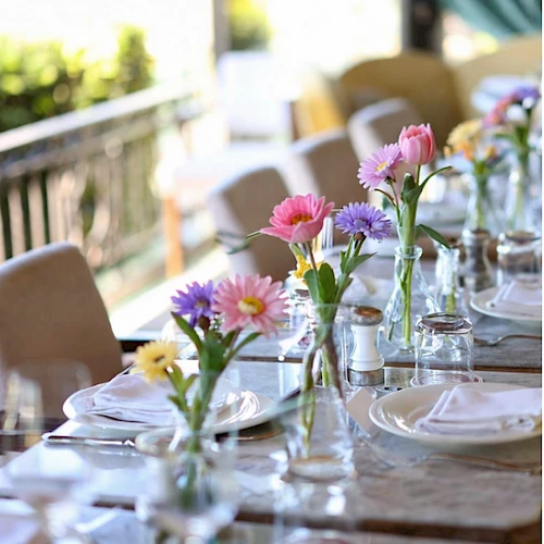 A long outdoor dining table adorned with glassware, pastel flowers in vases, and neatly set plates, suggesting a sunny garden meal.