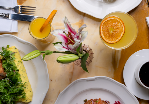 A bright brunch spread:を plates with greens, salmon toast, poached eggs on toast, orange juice, coffee, and a citrus salad, all on a marble table.