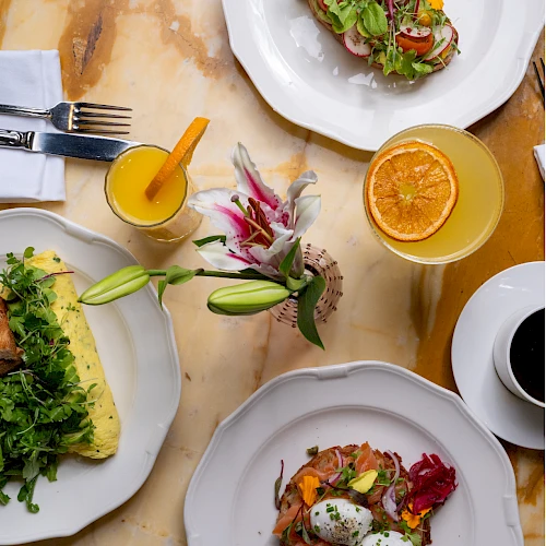A bright brunch spread:を plates with greens, salmon toast, poached eggs on toast, orange juice, coffee, and a citrus salad, all on a marble table.