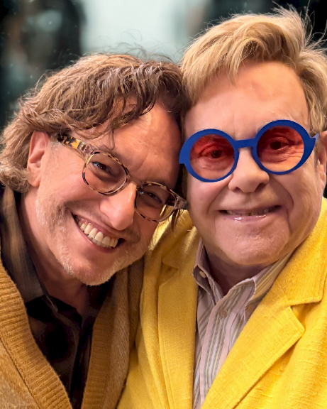 Two men smiling together, wearing bright yellow jackets; one with blue round glasses, posing closely.