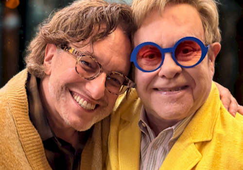 Two men smiling together, wearing bright yellow jackets; one with blue round glasses, posing closely.